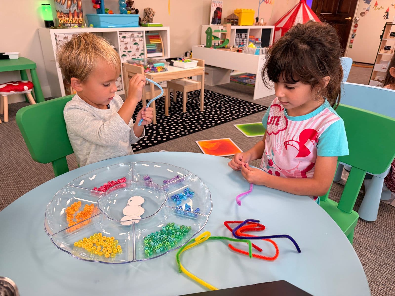Two preschoolers thread colorful beads onto pipe cleaners in a bright classroom.