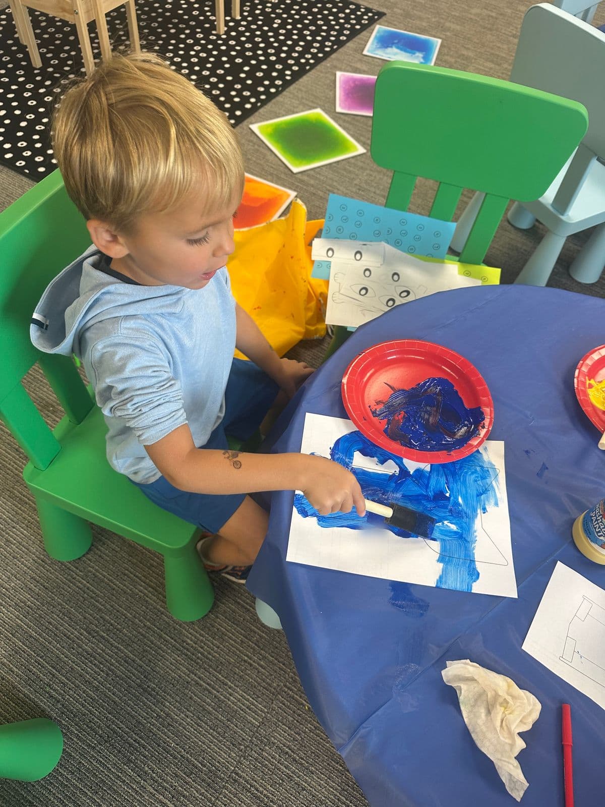 A boy paints bold blue strokes with a sponge brush at a craft table.