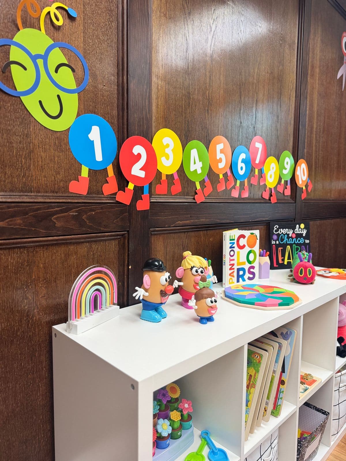A white shelf of toys, a colors book, and a learning sign beneath a paper caterpillar counting from 1 to 10 on a wood-paneled wall.