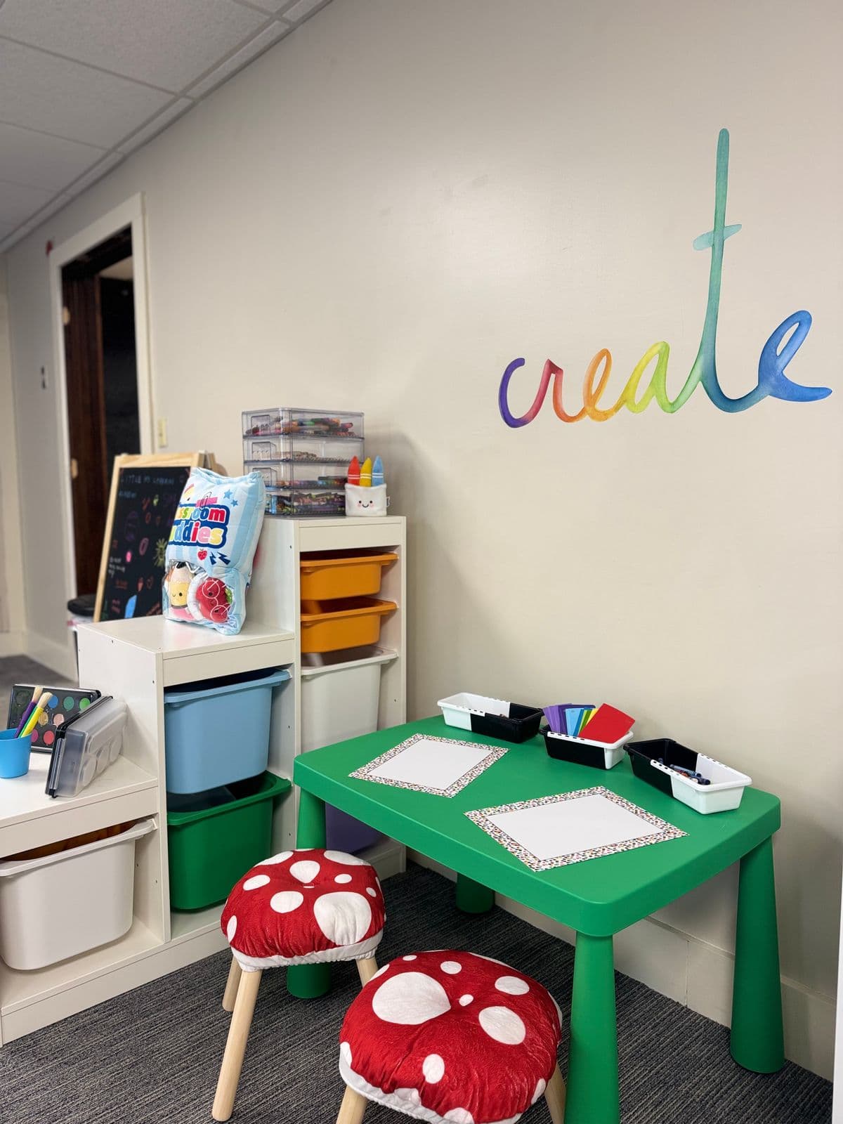 An art corner with a green table, drawing paper, crayon bins, and red mushroom stools beneath a rainbow “create” decal.