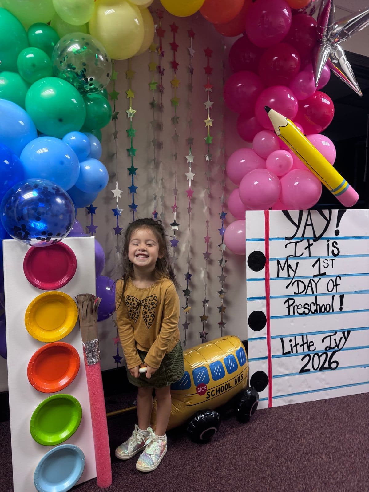A smiling preschooler stands under a rainbow balloon arch beside a sign celebrating her first day of preschool at Little Ivy.