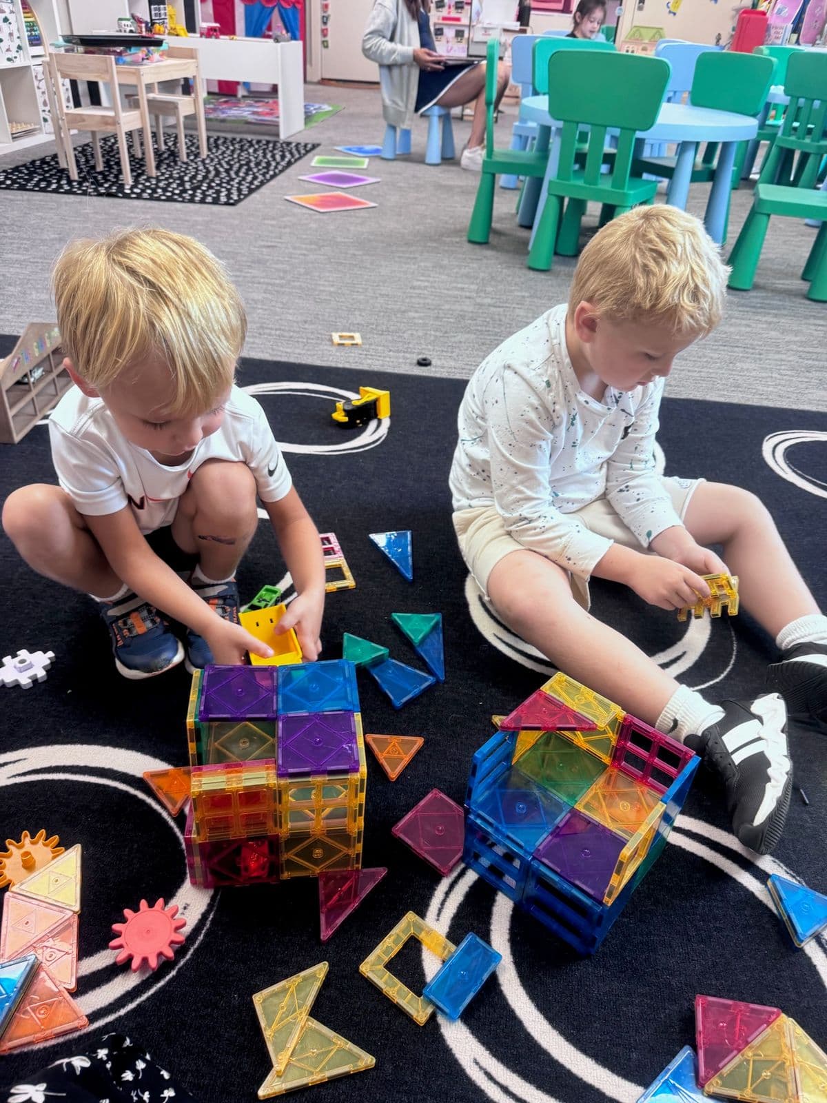 Two boys build colorful magnetic-tile structures on a patterned classroom rug.