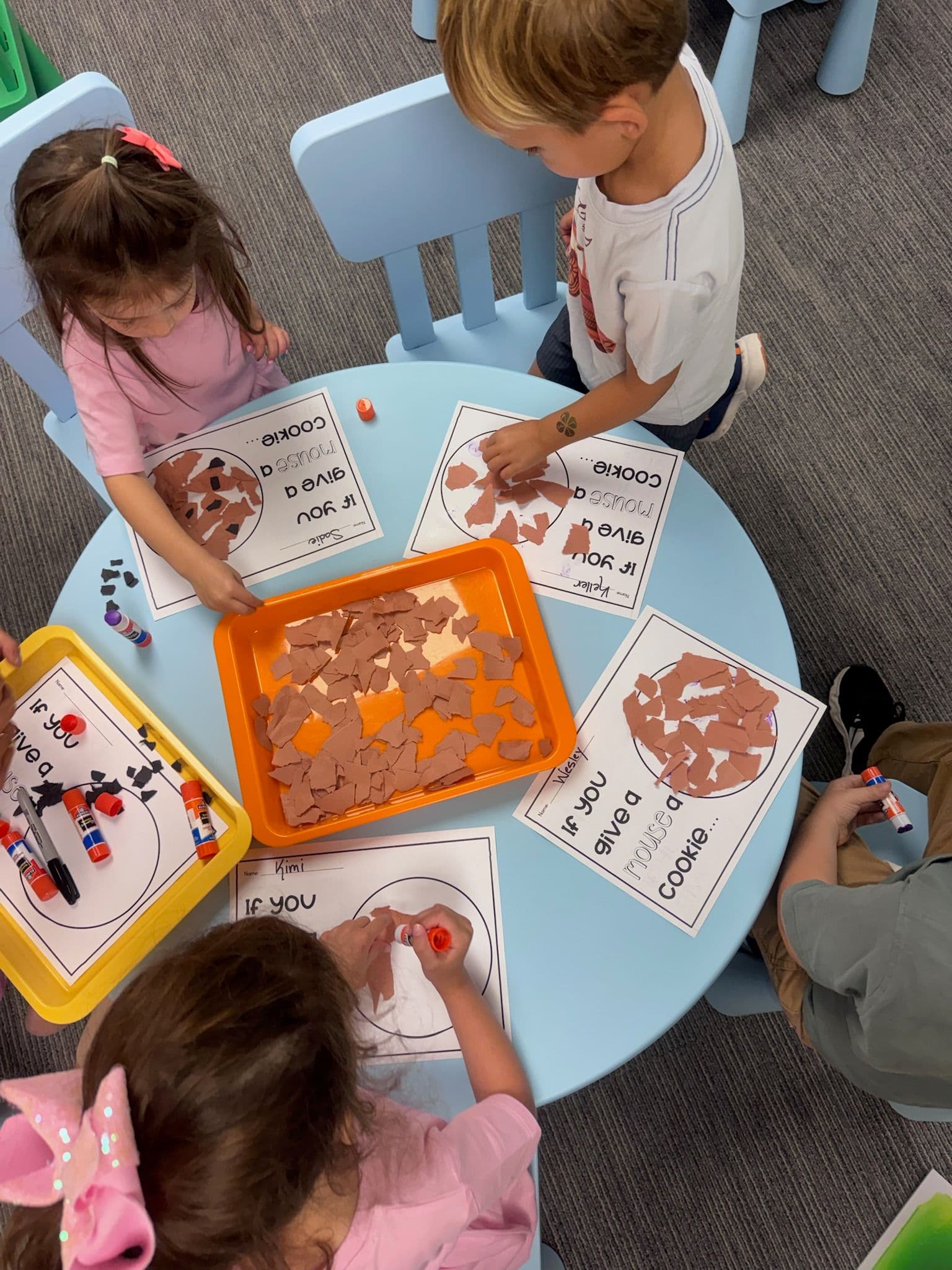 Preschoolers around a round blue table tear and glue brown paper “cookies” onto If You Give a Mouse a Cookie worksheets.