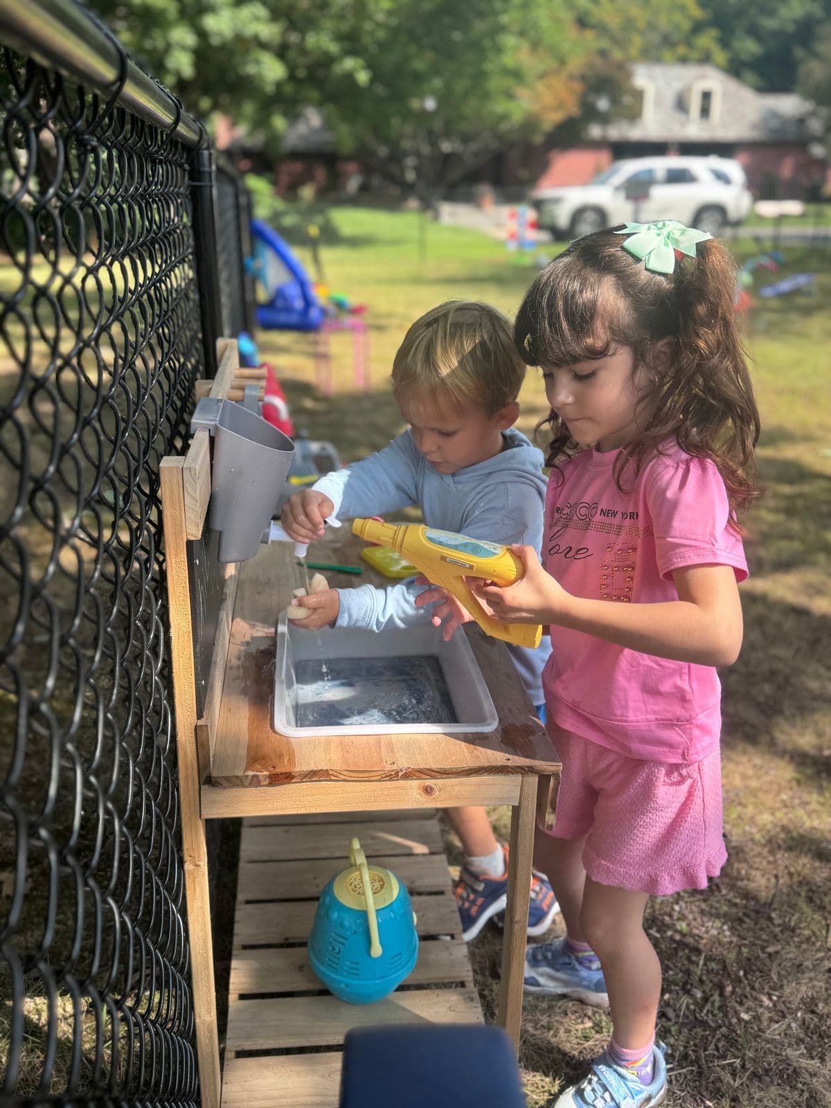 Two preschoolers pour and spray water at a wooden outdoor play sink beside a fence.