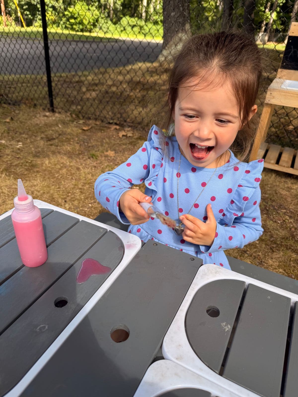 A laughing preschooler squeezes a dropper of pink liquid at an outdoor play table.