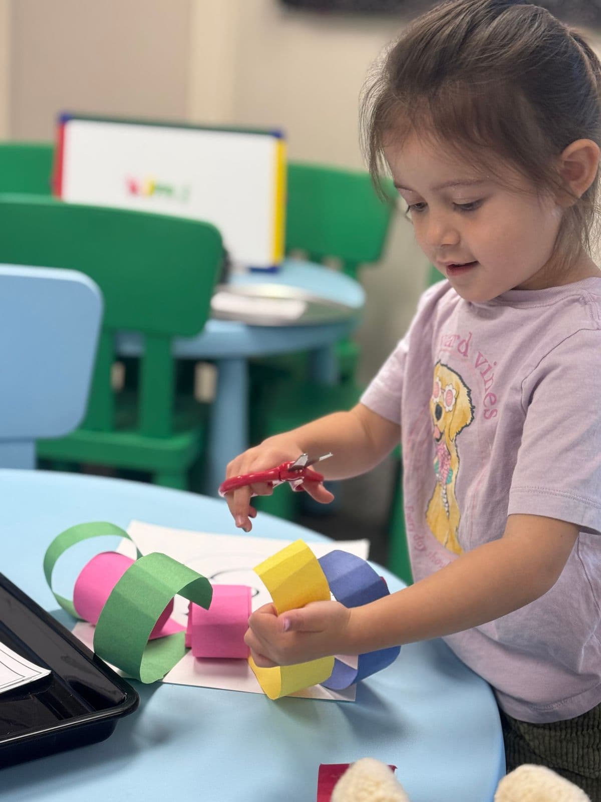 A preschooler holding safety scissors links colorful paper strips into a chain.