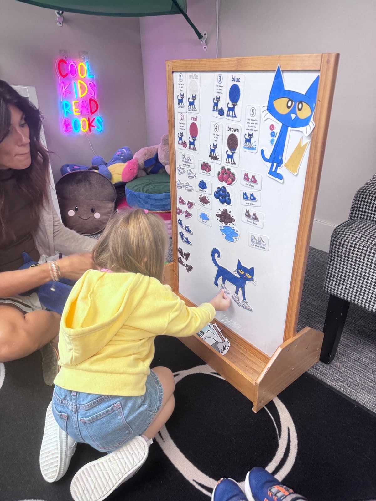A preschooler places story pieces on a Pete the Cat board while a teacher sits beside her in the reading nook.