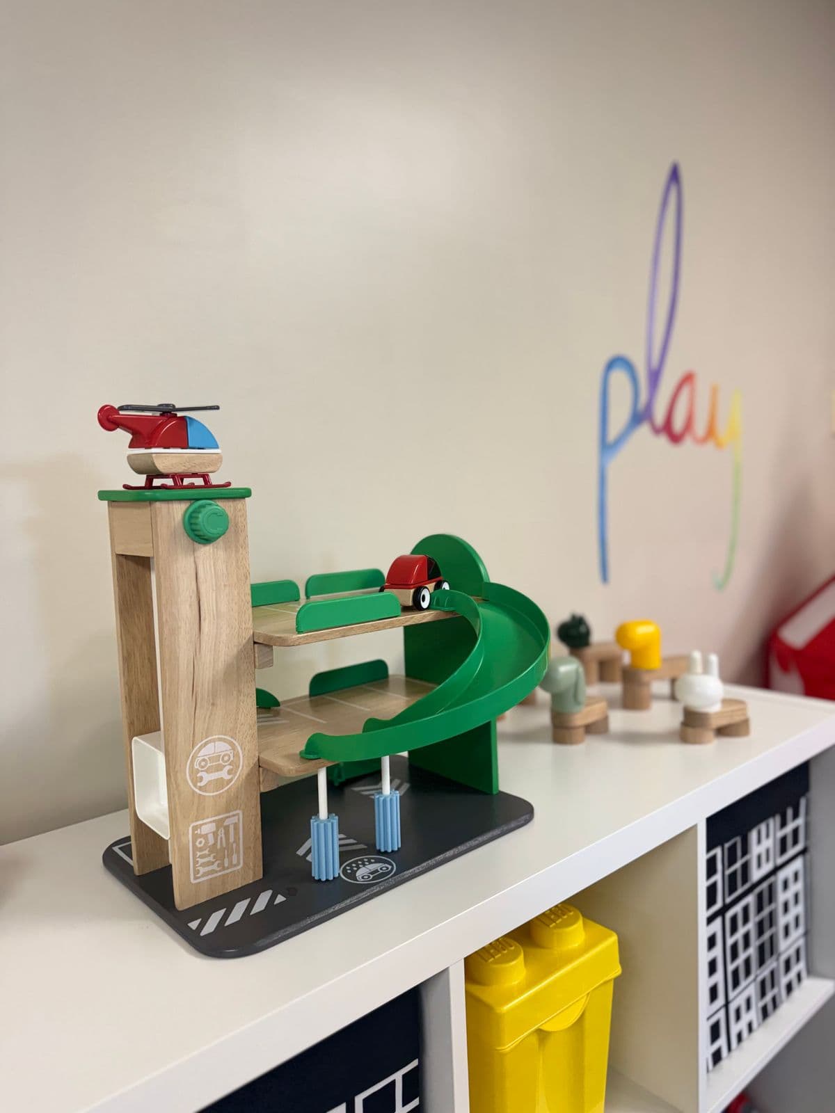 A wooden toy parking garage with a helicopter on top, on a white shelf beneath a rainbow “play” wall decal.