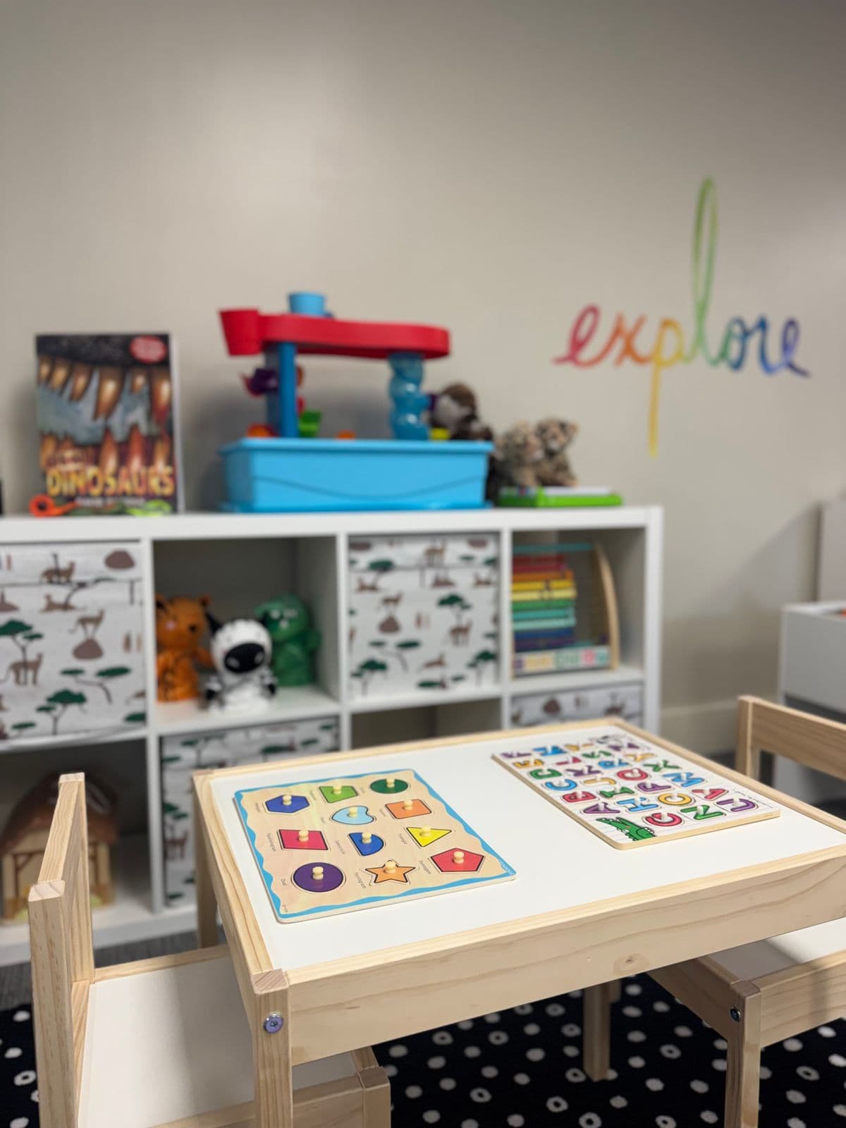 A child-size wooden table set with shape and alphabet peg puzzles in front of toy shelves.