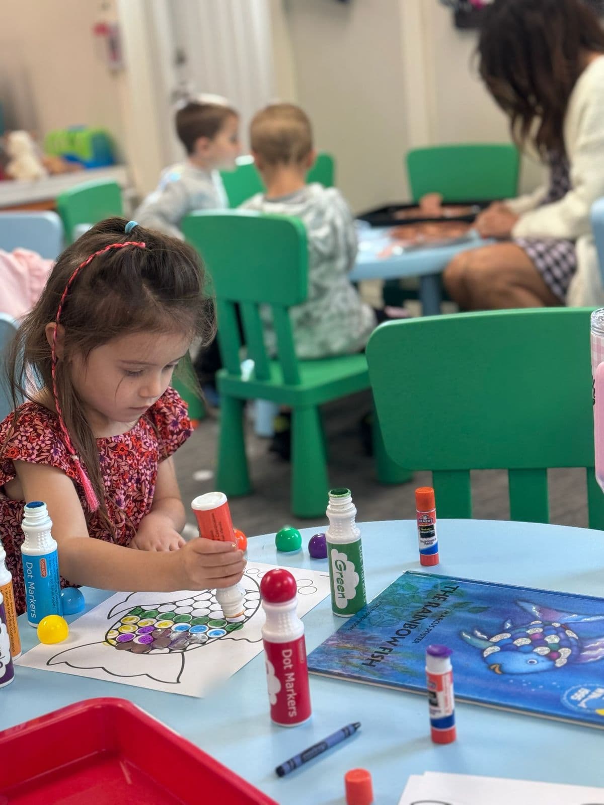 A preschooler decorates a Rainbow Fish worksheet with dot markers beside the storybook.