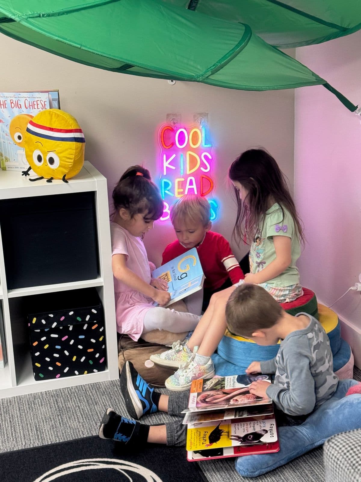 Four children share board books in a cozy reading nook beneath a glowing “Cool Kids Read Books” sign.
