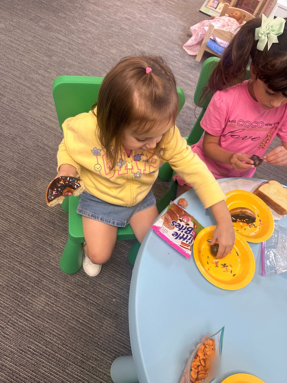 Two preschoolers enjoy a snack together at a round classroom table.
