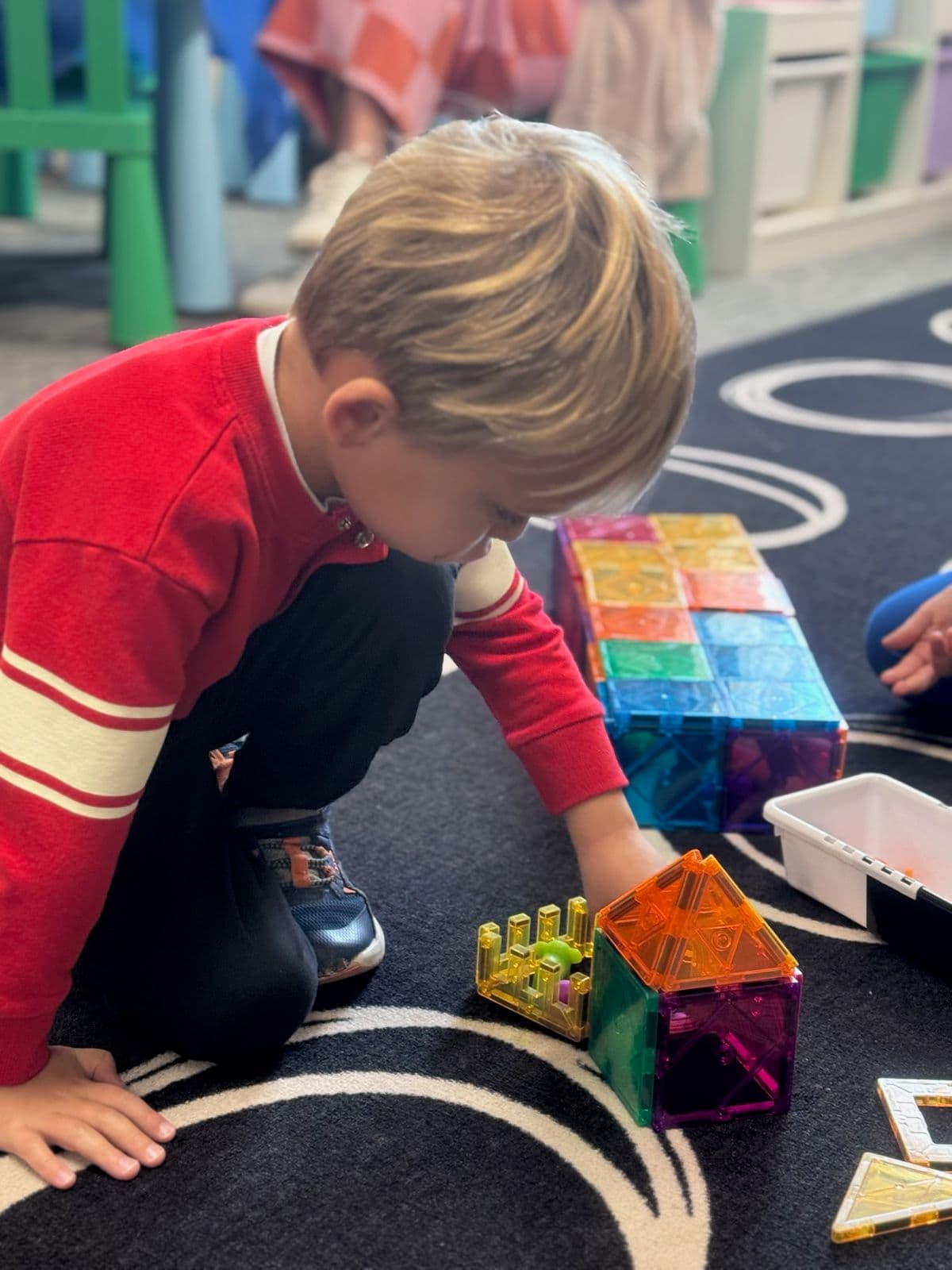 A boy builds a small house from colorful magnetic tiles on a classroom rug.