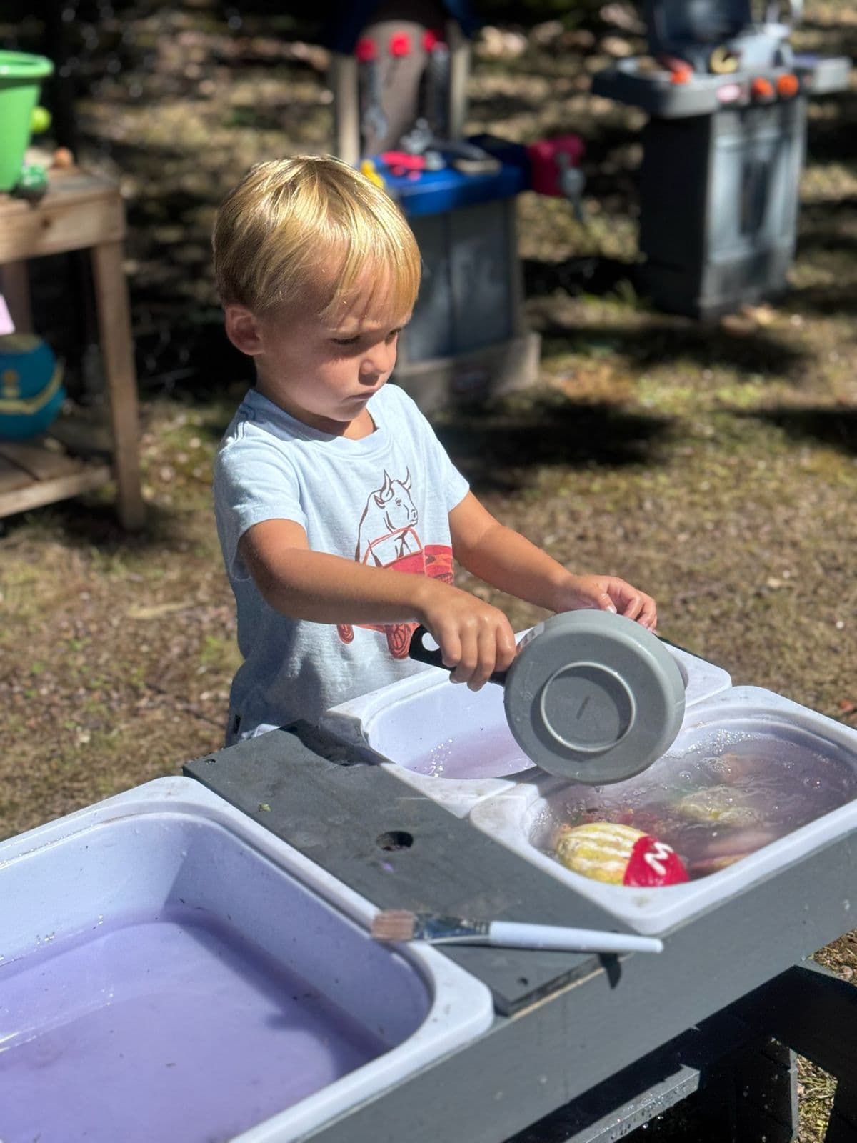 A boy pours water from a toy pot at an outdoor sensory water table.