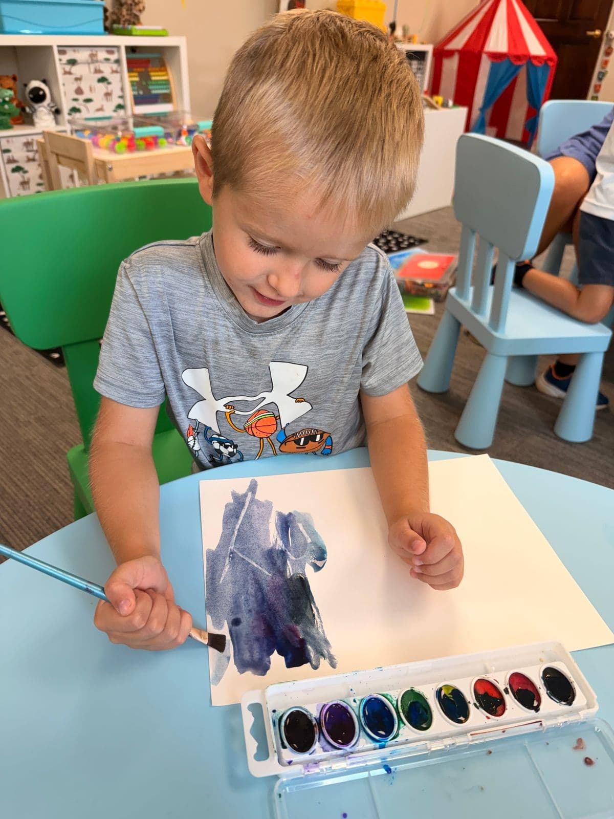 A boy brushes purple and blue watercolors onto paper at a classroom table.