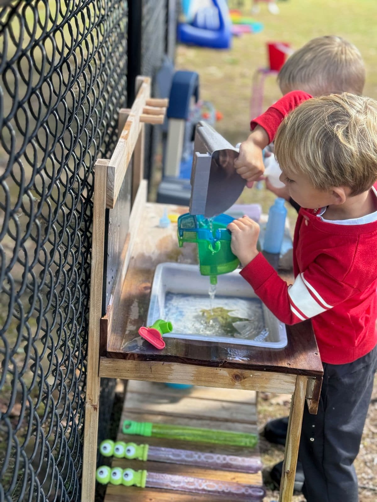 A boy pours water through a green watering can into an outdoor play sink.
