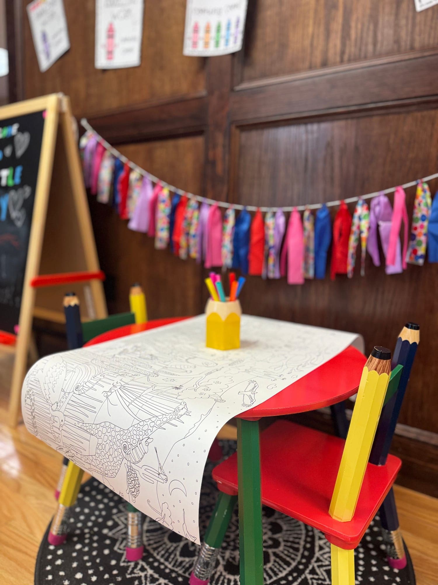 A red child-size table with pencil-shaped chairs, a coloring scroll, and a cup of markers beneath a ribbon garland.