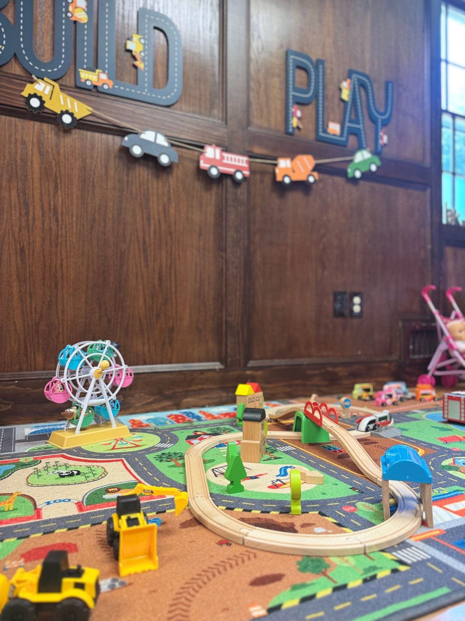 A wooden train set and toy construction trucks on a town play rug, beneath “Build” and “Play” letters on a wood-paneled wall.