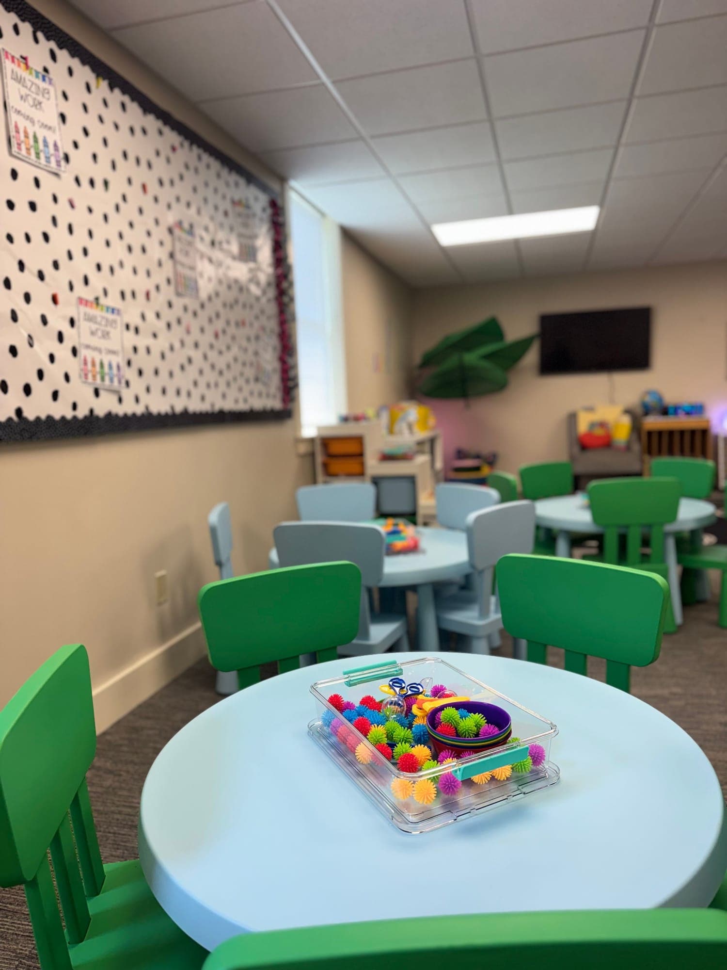 Round child-size tables with green and blue chairs, and a tub of sensory toys set out on the nearest one.