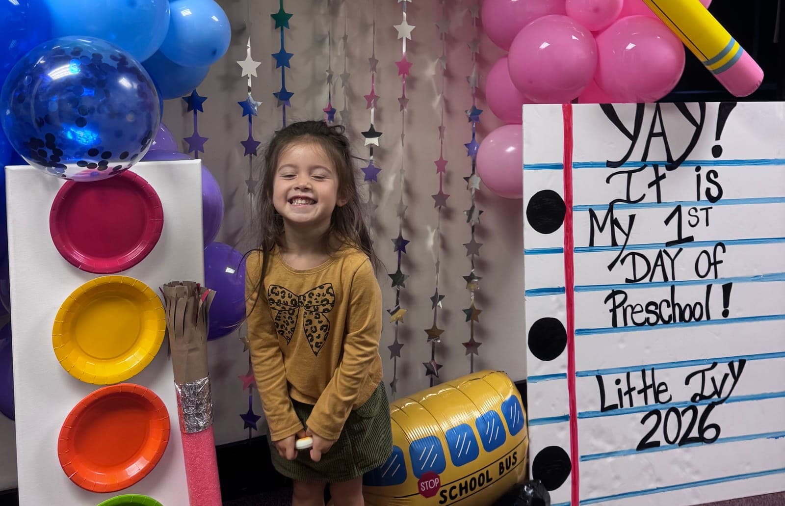 A beaming child on her first day at Little Ivy, beside a sign that reads “Yay! It is my 1st day of preschool!”