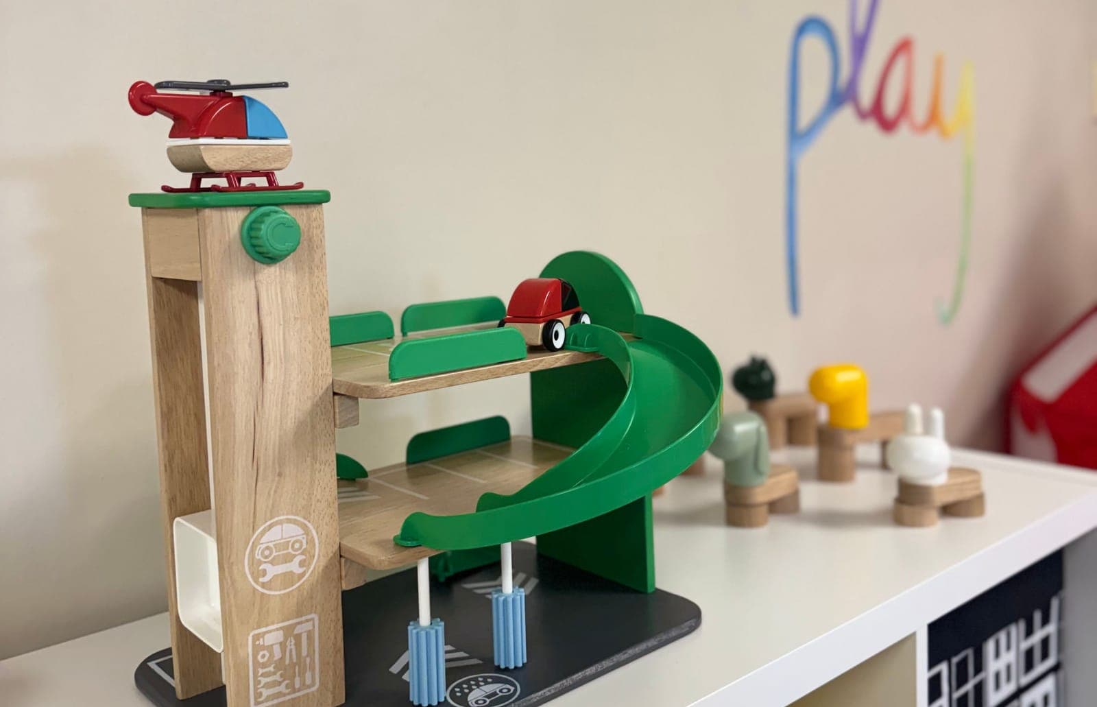 A wooden toy parking garage and play pieces on a classroom shelf, with the word “play” painted on the wall behind.