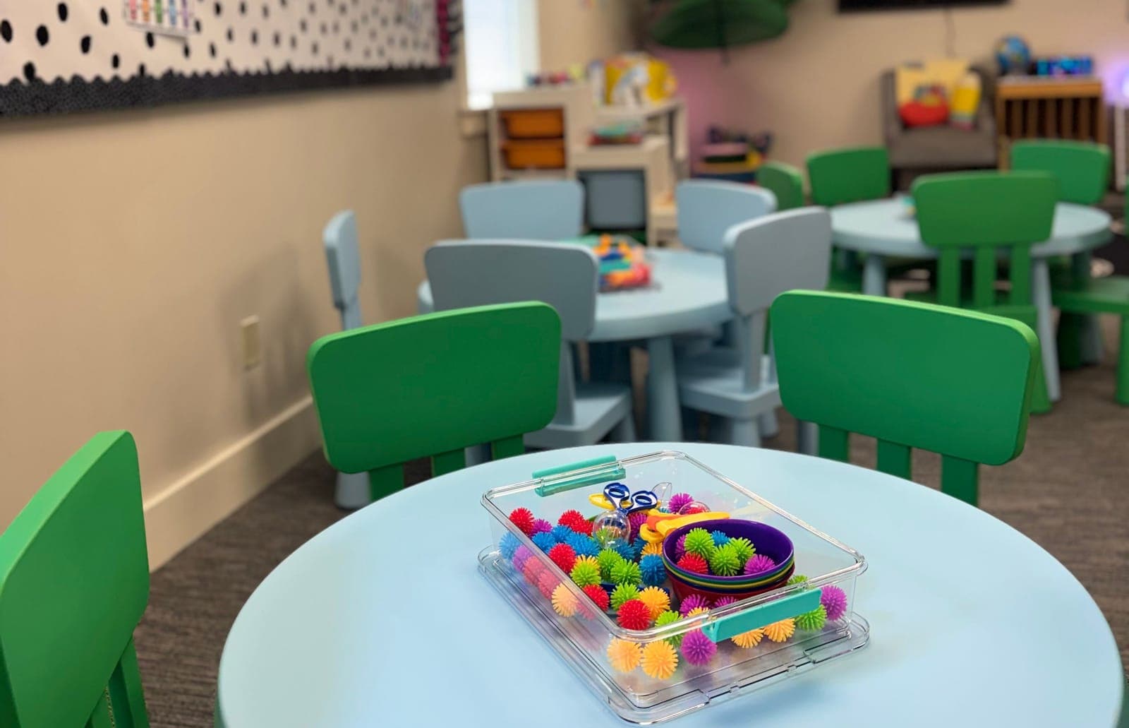 Round child-size tables and green chairs in a Little Ivy classroom, with a tub of sensory toys set out on the nearest table.